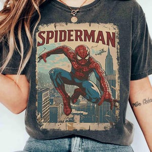 May include: Dark gray t-shirt featuring a vintage-style Spider-Man graphic. The design shows Spider-Man in a dynamic pose against a cityscape backdrop, with the word "SPIDERMAN" in bold letters. The shirt has a worn, distressed look.