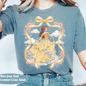May include: Blue-grey t-shirt with a cartoon illustration of a woman in a yellow gown, framed by a floral wreath and yellow ribbon. The text "Blue Jean Only With Comfort Color Adult" is at the bottom. A casual, comfortable top.