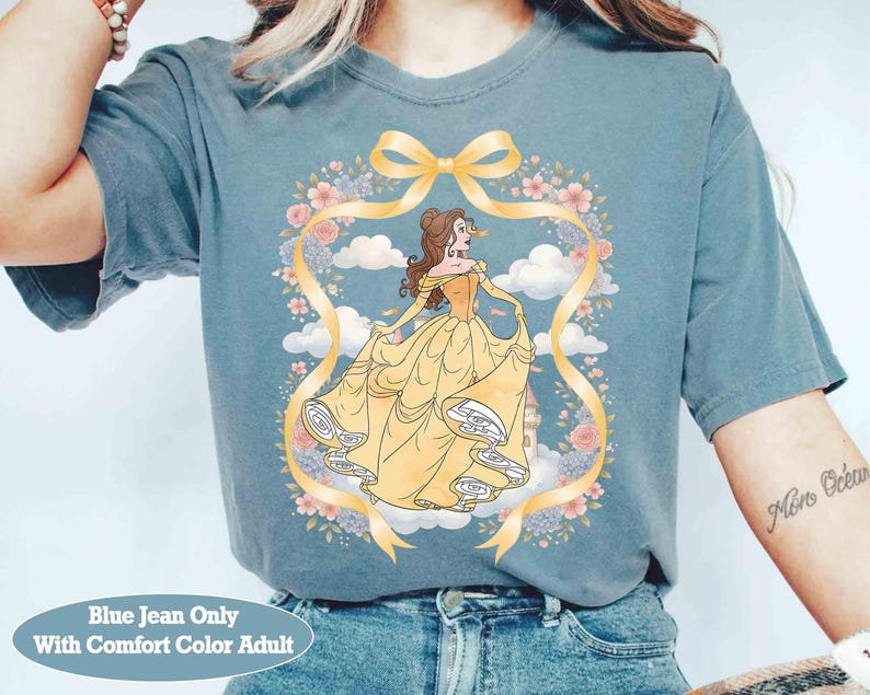 May include: Blue-grey t-shirt with a cartoon illustration of a woman in a yellow gown, framed by a floral wreath and yellow ribbon. The text "Blue Jean Only With Comfort Color Adult" is at the bottom. A casual, comfortable top.