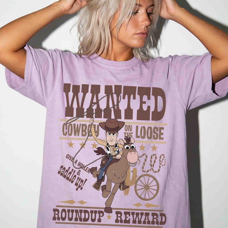 Pode incluir: Uma camiseta lil&aacute;s com um gr&aacute;fico de Woody de Toy Story montando um cavalo. A camisa tem as palavras "WANTED" e "COWBOY ON THE LOOSE" em letras marrons. Texto adicional inclui "GIVE A HOLLER & SADDLE UP!" e "ROUNDUP REWARD".