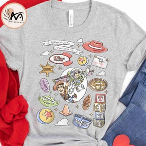May include: A light gray t-shirt featuring cartoon characters and phrases from the movie Toy Story. The design includes Woody, Buzz Lightyear, and other characters, along with the text "You've got a friend in me."