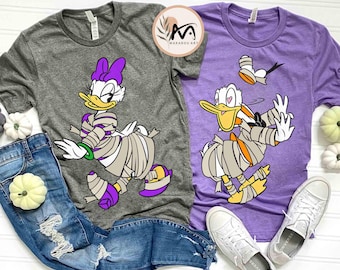 May include: Two t-shirts, one gray and one purple, featuring cartoon characters dressed as mummies. The gray shirt shows a duck with a purple bow, while the purple shirt shows a duck with a heart on its chest. Both shirts are Halloween-themed.
