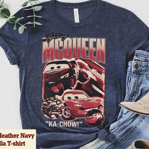 K&ouml;nnte beinhalten: Heather-Marineblaues Bella T-Shirt mit einem Aufdruck von Lightning McQueen aus dem Film Cars. Das Design enth&auml;lt den Text "Lightning McQueen" und "KA-CHOW!" in Rot und Wei&szlig;, mit Bildern des Autos.