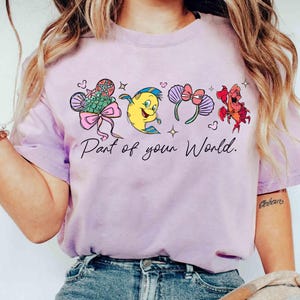 Puede incluir: Camiseta de color lila claro con ilustraciones de dibujos animados de personajes y accesorios coloridos. El texto "Part of your World" est&aacute; impreso en escritura negra debajo. La camiseta est&aacute; hecha de un material suave.
