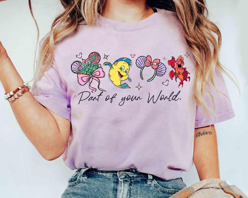 Puede incluir: Camiseta de color lila claro con ilustraciones de dibujos animados de personajes y accesorios coloridos. El texto "Part of your World" est&aacute; impreso en escritura negra debajo. La camiseta est&aacute; hecha de un material suave.