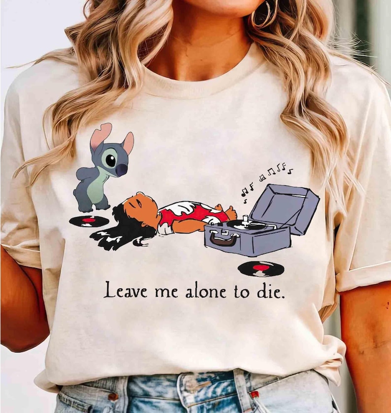 May include: Beige t-shirt featuring a cartoon illustration of Lilo and Stitch. Lilo is lying down near a record player, with Stitch standing nearby. The text "Leave me alone to die" is printed below the image.