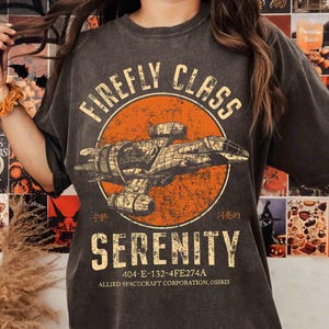 Puede incluir: Camiseta gris oscuro con un círculo naranja desgastado. El diseño presenta una ilustración de nave espacial y las palabras "FIREFLY CLASS SERENITY". Debajo, las palabras "404-E-132-4FE274A" y "ALLIED SPACECRAFT CORPORATION, OSIRIS".