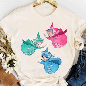 May include: Cream-colored t-shirt featuring a cartoon illustration of three fairies in teal, pink, and blue dresses and hats. The fairies are holding wands. The shirt is displayed with jeans and fall-themed decor.