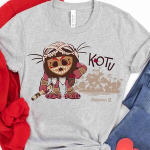Puede incluir: Camiseta gris claro con un personaje de dibujos animados con una m&aacute;scara de hueso y marcas tribales. La camiseta tiene las palabras "KOTU" y "MOANA 2" impresas. El personaje sostiene un bast&oacute;n.