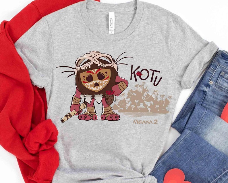Puede incluir: Camiseta gris claro con un personaje de dibujos animados con una m&aacute;scara de hueso y marcas tribales. La camiseta tiene las palabras "KOTU" y "MOANA 2" impresas. El personaje sostiene un bast&oacute;n.