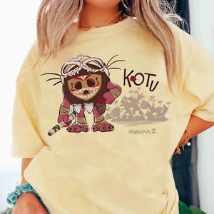 Puede incluir: Camiseta amarillo claro con un personaje de dibujos animados con una m&aacute;scara de calavera y dise&ntilde;os tribales. El personaje sostiene un bast&oacute;n, con una escena de isla tropical de fondo. La palabra "KOTU" est&aacute; encima del personaje, con "MOANA 2" debajo.