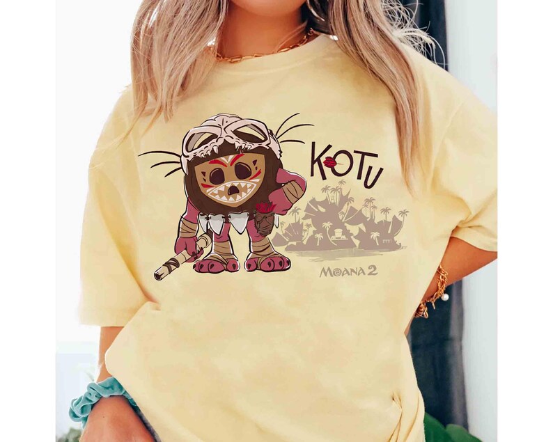 Puede incluir: Camiseta amarillo claro con un personaje de dibujos animados con una m&aacute;scara de calavera y dise&ntilde;os tribales. El personaje sostiene un bast&oacute;n, con una escena de isla tropical de fondo. La palabra "KOTU" est&aacute; encima del personaje, con "MOANA 2" debajo.