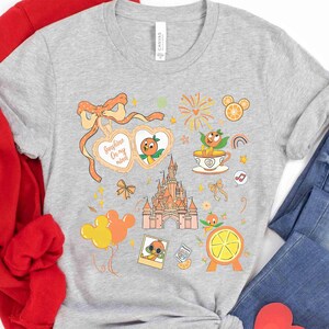 May include: A gray t-shirt featuring a colorful graphic design with orange and yellow elements. The design includes a castle, orange characters, Mickey Mouse balloons, and the text "Sunshine On my mind".