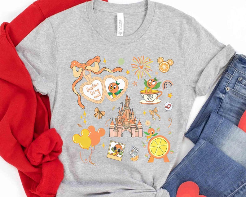 May include: A gray t-shirt featuring a colorful graphic design with orange and yellow elements. The design includes a castle, orange characters, Mickey Mouse balloons, and the text "Sunshine On my mind".