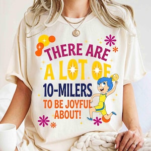 Könnte beinhalten: Cremefarbenes T-Shirt mit dem Text "THERE ARE A LOT OF 10-MILERS TO BE JOYFUL ABOUT!" in verschiedenen Farben. Das Shirt zeigt eine Cartoonfigur in einem gelb-blauen Outfit.
