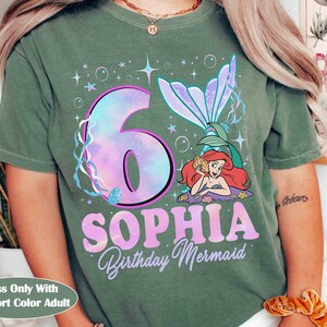 Puede incluir: Camiseta verde con un dise&ntilde;o de sirena. El dise&ntilde;o incluye el n&uacute;mero 6 en un estilo tie-dye, el nombre "Sophia" y las palabras "Birthday Mermaid". Tambi&eacute;n aparece una sirena de dibujos animados.