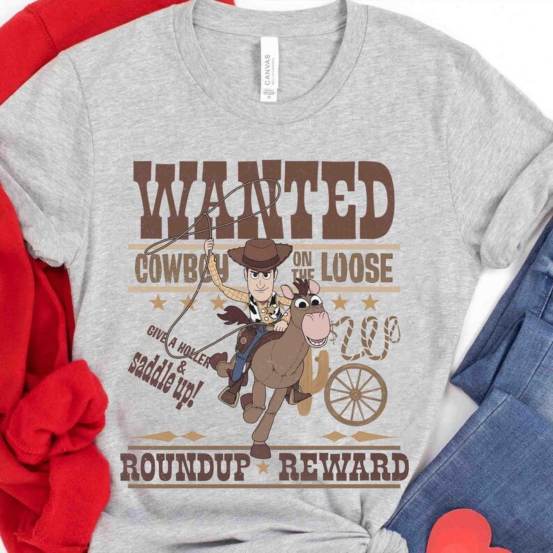 Pode incluir: T-shirt cinzenta clara com um cowboy de desenho animado montado num cavalo e o texto castanho "WANTED COWBOY ON THE LOOSE". A camisa tamb&eacute;m inclui as frases "Give a holler & saddle up!" e "Roundup Reward".