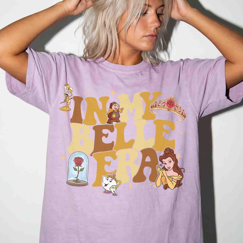 Op de afbeelding: Een lila T-shirt met de tekst "IN MY BELLE ERA" in bruine en gele retro letters. Het shirt heeft cartoon illustraties van personages en objecten uit Belle en het Beest.