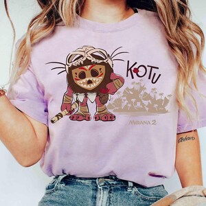 Puede incluir: Camiseta de color lila claro con un personaje de dibujos animados con una m&aacute;scara y un bast&oacute;n. La palabra "KOTU" est&aacute; encima del personaje y "MOANA 2" debajo. El personaje es marr&oacute;n, rojo y beige.
