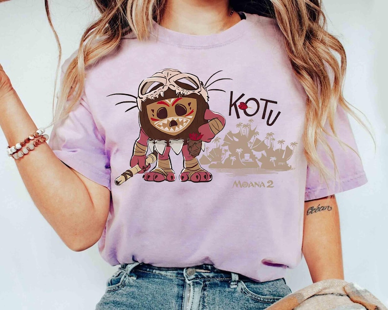 Puede incluir: Camiseta de color lila claro con un personaje de dibujos animados con una m&aacute;scara y un bast&oacute;n. La palabra "KOTU" est&aacute; encima del personaje y "MOANA 2" debajo. El personaje es marr&oacute;n, rojo y beige.