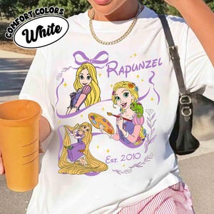 K&ouml;nnte beinhalten: Wei&szlig;es T-Shirt mit einem Rapunzel-Motiv. Das Design zeigt Illustrationen von Rapunzel mit langen blonden Haaren, einem Cham&auml;leon und einer Farbpalette. Der Text "Rapunzel" und "Est. 2010" sind ebenfalls enthalten.