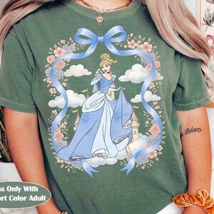 May include: Moss green Comfort Color adult t-shirt with a Cinderella graphic. The design features a blue ribbon, floral wreath, and a cartoon image of Cinderella in a blue gown. The text "Moss Only With Comfort Color Adult" is at the bottom.
