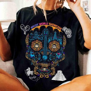 May include: Black t-shirt featuring a colorful sugar skull design with a guitar, flowers, and the text "EPCOT WORLD SHOWCASE". The design includes images of a llama, maracas, and a pyramid, with the word "Mexico" below.