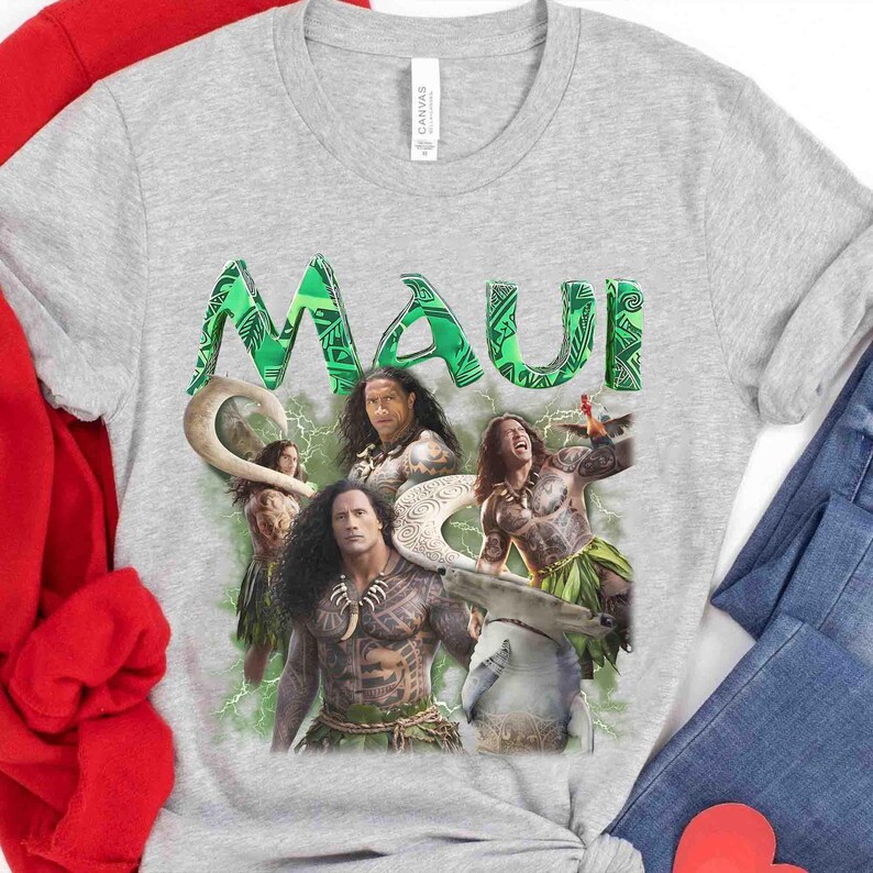 May include: A heather gray t-shirt featuring a graphic print of the character Maui from the movie Moana. The design includes the word MAUI in large green patterned letters above images of the character in various poses with a hook.