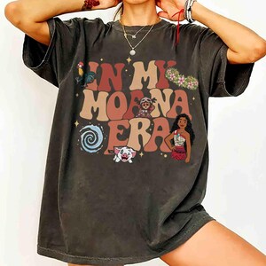 Op de afbeelding: Donkergrijs t-shirt met de tekst "IN MY MOANA ERA" in bruine, oranje en roze letters. Het shirt heeft cartoon illustraties van personages en elementen uit de film Moana, waaronder een haan, een varken en Moana zelf.