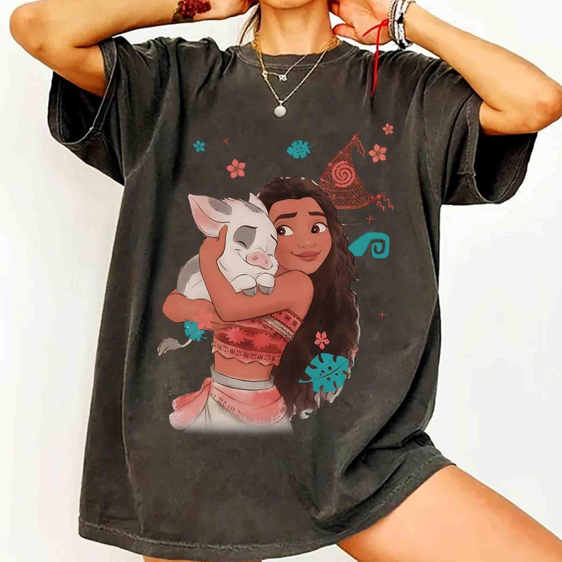 Puede incluir: Una camiseta gris carb&oacute;n de manga corta con un estampado gr&aacute;fico de un personaje de pelo largo abrazando a un peque&ntilde;o lech&oacute;n. El dise&ntilde;o incluye peque&ntilde;os patrones florales y tribales en tonos rojos, rosas y azul cerceta alrededor de las figuras.