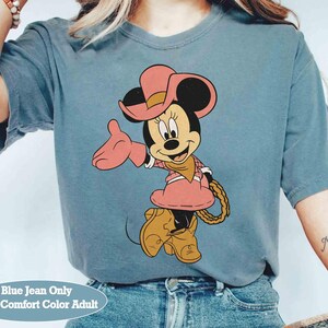 Puede incluir: Camiseta azul vaquero con Minnie Mouse de dibujos animados vestida de vaquera. Minnie lleva sombrero, vestido y botas rosas, con una cuerda en la cintura. El texto "Blue Jean Only With Comfort Color Adult" est&aacute; en la parte inferior.