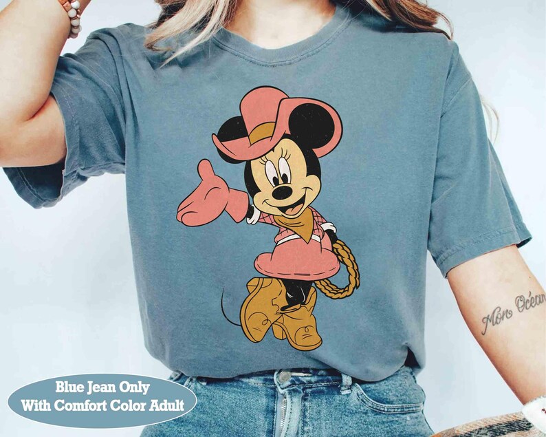 Puede incluir: Camiseta azul vaquero con Minnie Mouse de dibujos animados vestida de vaquera. Minnie lleva sombrero, vestido y botas rosas, con una cuerda en la cintura. El texto "Blue Jean Only With Comfort Color Adult" est&aacute; en la parte inferior.