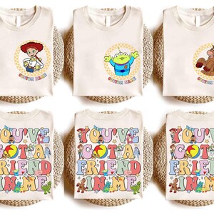Op de afbeelding: Cr&egrave;mekleurige T-shirts met stripfiguren en tekst uit de film Toy Story. Ontwerpen omvatten Jessie, een alien en Bullseye in cirkelvormige frames. Andere shirts tonen de tekst "YOU'VE GOT A FRIEND IN ME" in kleurrijke letters.
