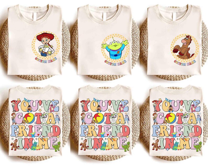 Op de afbeelding: Cr&egrave;mekleurige T-shirts met stripfiguren en tekst uit de film Toy Story. Ontwerpen omvatten Jessie, een alien en Bullseye in cirkelvormige frames. Andere shirts tonen de tekst "YOU'VE GOT A FRIEND IN ME" in kleurrijke letters.