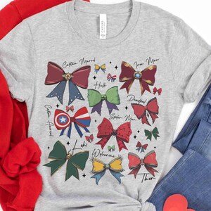 May include: A light gray t-shirt featuring colorful bow designs representing various Marvel characters, including Captain Marvel, Iron Man, and Thor. The names of the characters are written above each bow. The shirt is paired with blue jeans and a red sweater.