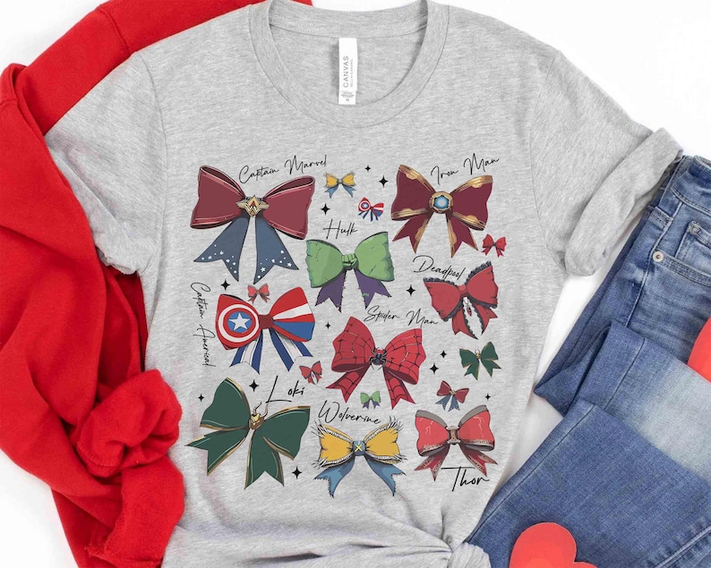 May include: A light gray t-shirt featuring colorful bow designs representing various Marvel characters, including Captain Marvel, Iron Man, and Thor. The names of the characters are written above each bow. The shirt is paired with blue jeans and a red sweater.