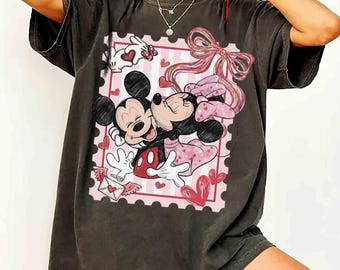 May include: Dark gray t-shirt featuring a graphic of Mickey and Minnie Mouse embracing within a pink postage stamp design. The design includes hearts, a bow, and a love letter, creating a romantic theme.