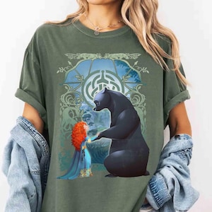 Puede incluir: Camiseta verde oliva con un gráfico de una niña pelirroja con un vestido azul y un oso negro. El diseño está ambientado sobre un fondo de una ventana azul y verde con detalles de nudos celtas. La camiseta tiene las mangas dobladas.