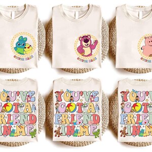 Op de afbeelding: Cr&egrave;mekleurige T-shirts met stripfiguren en de tekst "CUSTOM NAME" in een cirkel. De onderste rij shirts toont de zin "YOU'VE GOT A FRIEND IN ME" in kleurrijke letters met stripfiguren.