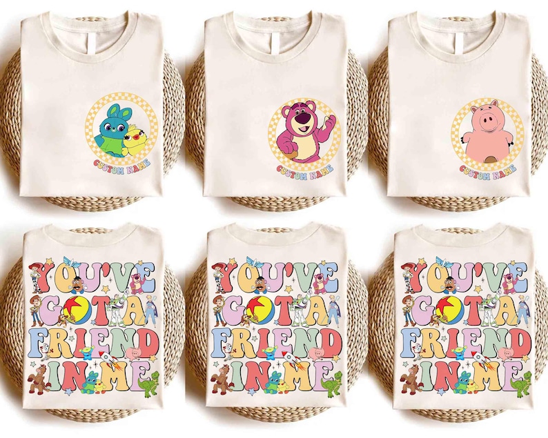 Op de afbeelding: Cr&egrave;mekleurige T-shirts met stripfiguren en de tekst "CUSTOM NAME" in een cirkel. De onderste rij shirts toont de zin "YOU'VE GOT A FRIEND IN ME" in kleurrijke letters met stripfiguren.