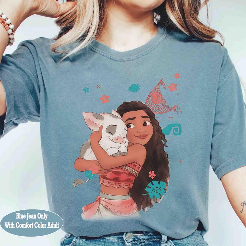 Puede incluir: Una camiseta de color azul vaquero con una ilustraci&oacute;n gr&aacute;fica de un personaje de pelo largo abrazando a un peque&ntilde;o lech&oacute;n. El dise&ntilde;o incluye peque&ntilde;os patrones florales y tribales en tonos rosa, naranja y turquesa alrededor.