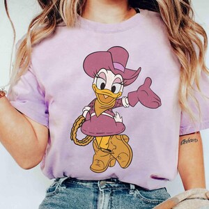 Puede incluir: Camiseta lila claro con una ilustraci&oacute;n de dibujos animados de Daisy Duck vestida de vaquera. Daisy lleva un sombrero, guantes y botas moradas, y sostiene una cuerda. El dise&ntilde;o est&aacute; centrado en la camiseta.
