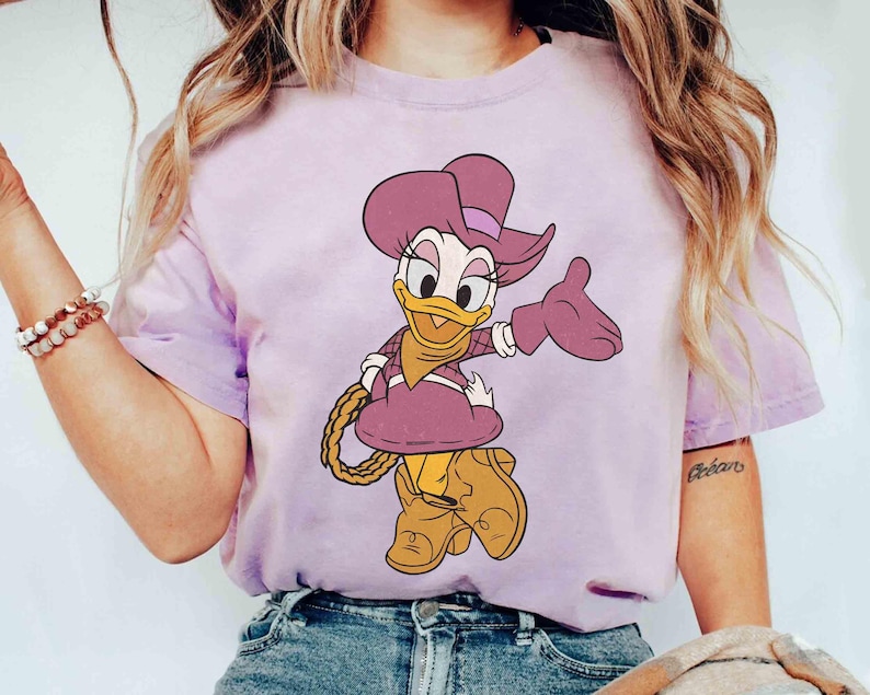 Puede incluir: Camiseta lila claro con una ilustraci&oacute;n de dibujos animados de Daisy Duck vestida de vaquera. Daisy lleva un sombrero, guantes y botas moradas, y sostiene una cuerda. El dise&ntilde;o est&aacute; centrado en la camiseta.