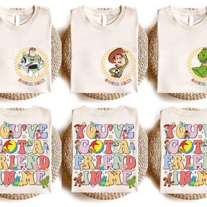 Op de afbeelding: Cr&egrave;mekleurige T-shirts met de stripfiguren Buzz Lightyear, Woody en Rex, elk in een cirkelvormig ontwerp. De onderste rij toont shirts met de tekst "YOU'VE GOT A FRIEND IN ME" in kleurrijke retro letters, met verschillende Toy Story-personages.
