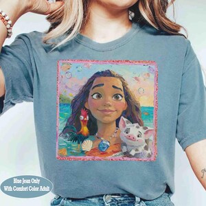 Puede incluir: Una camiseta azul gris&aacute;ceo con un estampado gr&aacute;fico cuadrado de un personaje de dibujos animados con cabello largo casta&ntilde;o, un gallo y un cerdito. El dise&ntilde;o incluye un fondo de playa con burbujas y un borde rosa. Cuello redondo y mangas cortas.