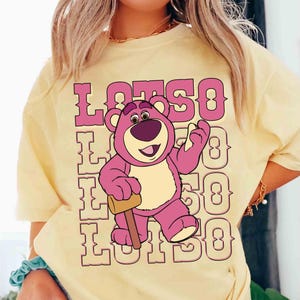 May include: A light yellow t-shirt featuring a graphic of Lotso, the pink bear from Toy Story, holding a mallet. The word "LOTSO" is printed in pink above and below the bear.