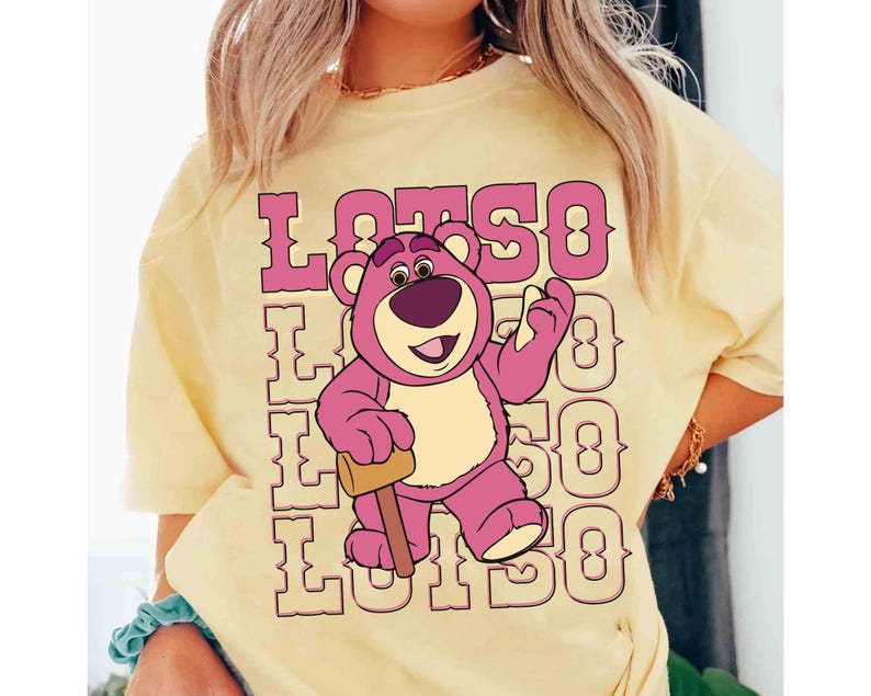 May include: A light yellow t-shirt featuring a graphic of Lotso, the pink bear from Toy Story, holding a mallet. The word "LOTSO" is printed in pink above and below the bear.