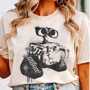 Könnte beinhalten: Beiges T-Shirt mit einer schwarz-weißen Grafik des Roboters WALL-E. Der Roboter hat große, fernglasartige Augen und Panzerketten. Das Wort "WALL" ist auf dem Körper des Roboters zu sehen. Das Design hat einen Used-Look im Sprühstil.
