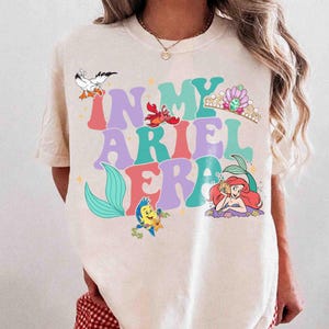 Op de afbeelding: Cr&egrave;mekleurig T-shirt met de tekst "IN MY ARIEL ERA" in kleurrijke letters. Het ontwerp bevat illustraties van Ariel, Botje, Sebastiaan, een zeemeeuw en een kroon, in een grillige cartoonstijl.