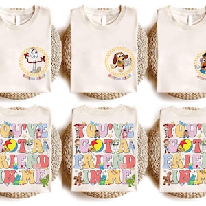 Op de afbeelding: Zes cr&egrave;mekleurige t-shirts met kleurrijke graphics. De bovenste rij heeft karakterontwerpen in cirkels, terwijl de onderste rij de tekst "YOU'VE GOT A FRIEND IN ME" in een retro lettertype weergeeft, met verschillende karakterillustraties.